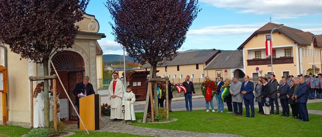Eine Gruppe von Menschen versammelt sich vor einer Kirche, wobei zwei Priester an einem Podium stehen. Die Umgebung umfasst Bäume, einen klaren blauen Himmel und eine kleine Menschenmenge.