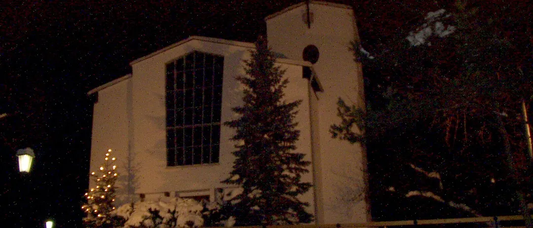 Ein weißes Kirchengebäude ist nachts beleuchtet, mit einem Weihnachtsbaum davor und Schnee auf dem Boden.