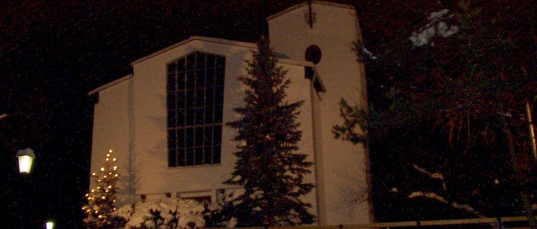 Ein weißes Kirchengebäude ist nachts beleuchtet, mit einem Weihnachtsbaum davor und Schnee auf dem Boden.