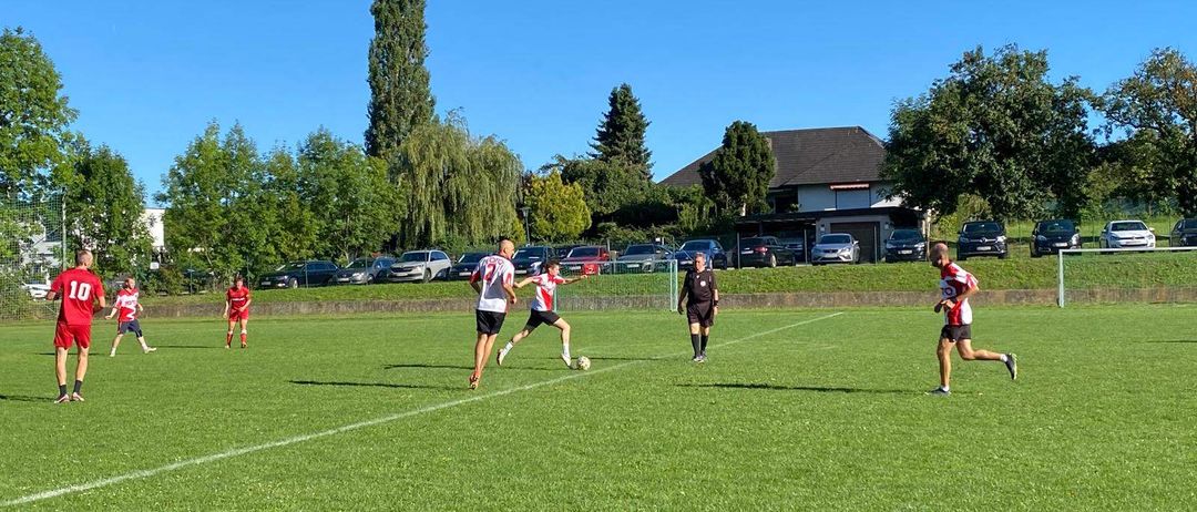 Ein Fußballspiel ist auf einem Rasenfeld im Gange, mit Spielern in roten und weißen Trikots. Ein Spieler läuft auf den Ball zu, während andere auf dem Feld positioniert sind. Im Hintergrund befinden sich Bäume und geparkte Autos in der Nähe eines Hauses.