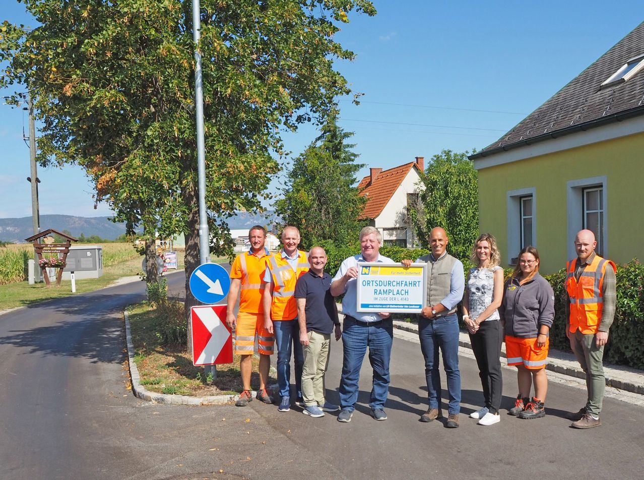Eine Gruppe von Personen in Sicherheitsausrüstung steht vor einem gelben Haus mit einem Schild, das Ortsdurchfahrt Ramplach besagt. Sie halten ein großes blaues Schild mit weißer Schrift.