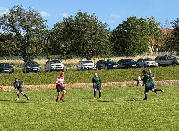 Eine Gruppe von Fußballspielern spielt auf einem Rasenfeld, an dessen Seite einige Autos geparkt sind.