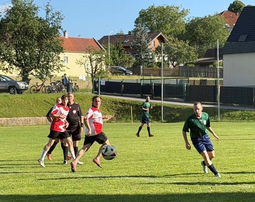 Eine Gruppe von Fußballspielern in roten und weißen Trikots jagt einem Ball auf einem Rasenfeld nach, ein Schiedsrichter in Schwarz ist in der Nähe. Im Hintergrund sind Häuser und geparkte Autos zu sehen.
