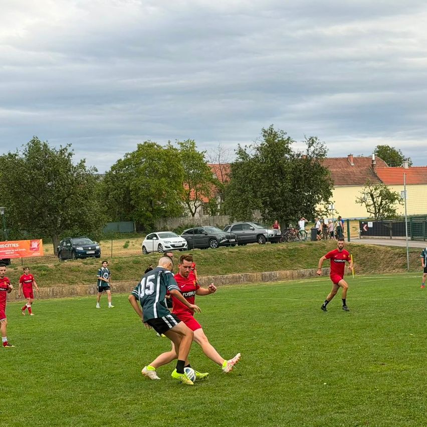 Eine Gruppe von Fußballspielern in roten Trikots spielt auf einem Rasenfeld. Einige Spieler rennen auf den Ball zu, während andere stehen. Im Hintergrund sind geparkte Autos und ein Gebäude mit einem roten Dach zu sehen.