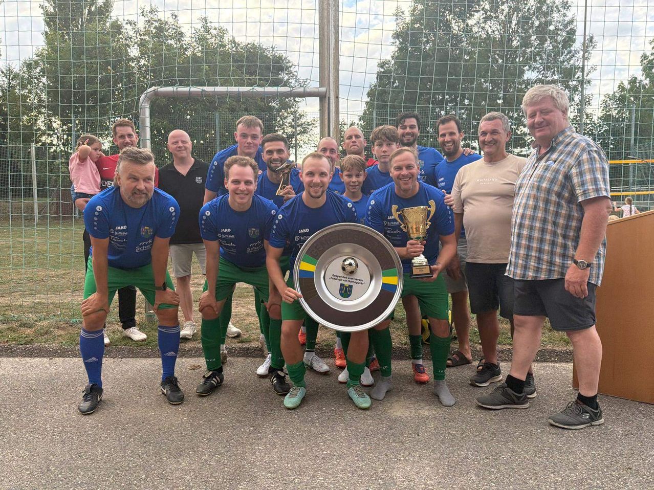 Eine Fußballmannschaft in blauen und grünen Trikots posiert mit einem Pokal und einem großen silbernen Schild vor einem Tornetz.