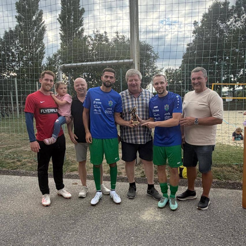 Eine Gruppe von Männern und einem Kind steht vor einem Fußballtornetz, lächelt und hält eine Trophäe.