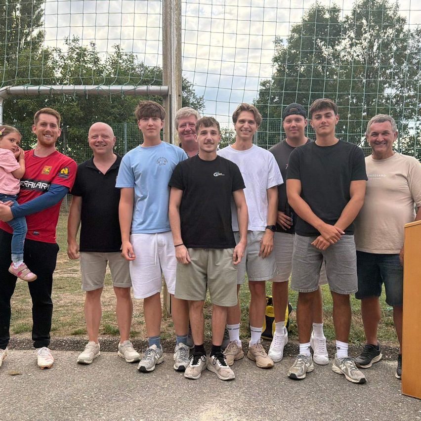 Eine Gruppe von Männern und einem kleinen Mädchen steht vor einem Fußballtornetz und lächelt für ein Foto. Der Hintergrund zeigt einen Rasenbereich mit Bäumen.