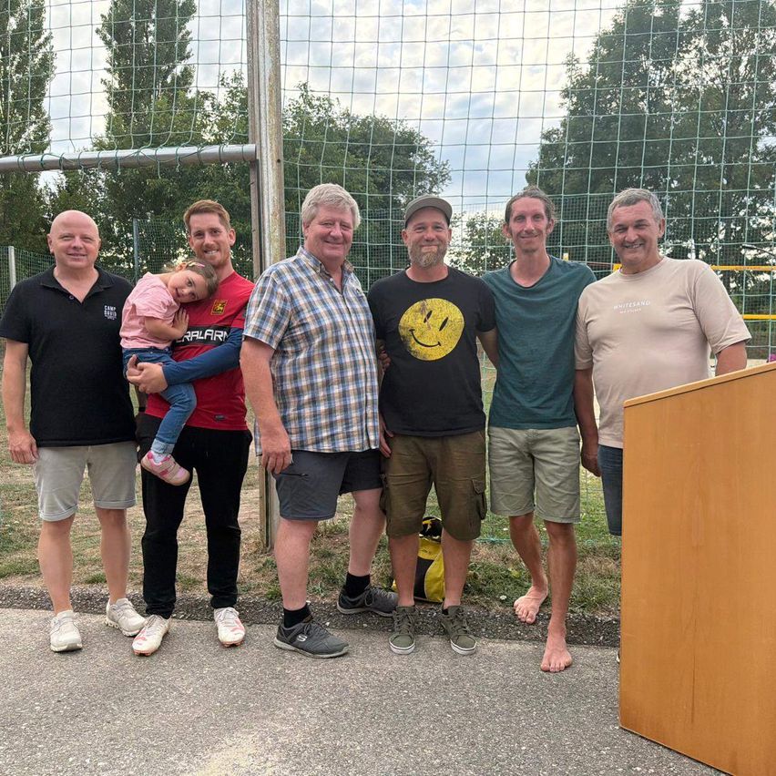 Sechs Männer und ein junges Mädchen stehen vor einem Fußballnetz und lächeln für ein Foto. Der Mann auf der linken Seite hält das Mädchen, das Pink trägt.
