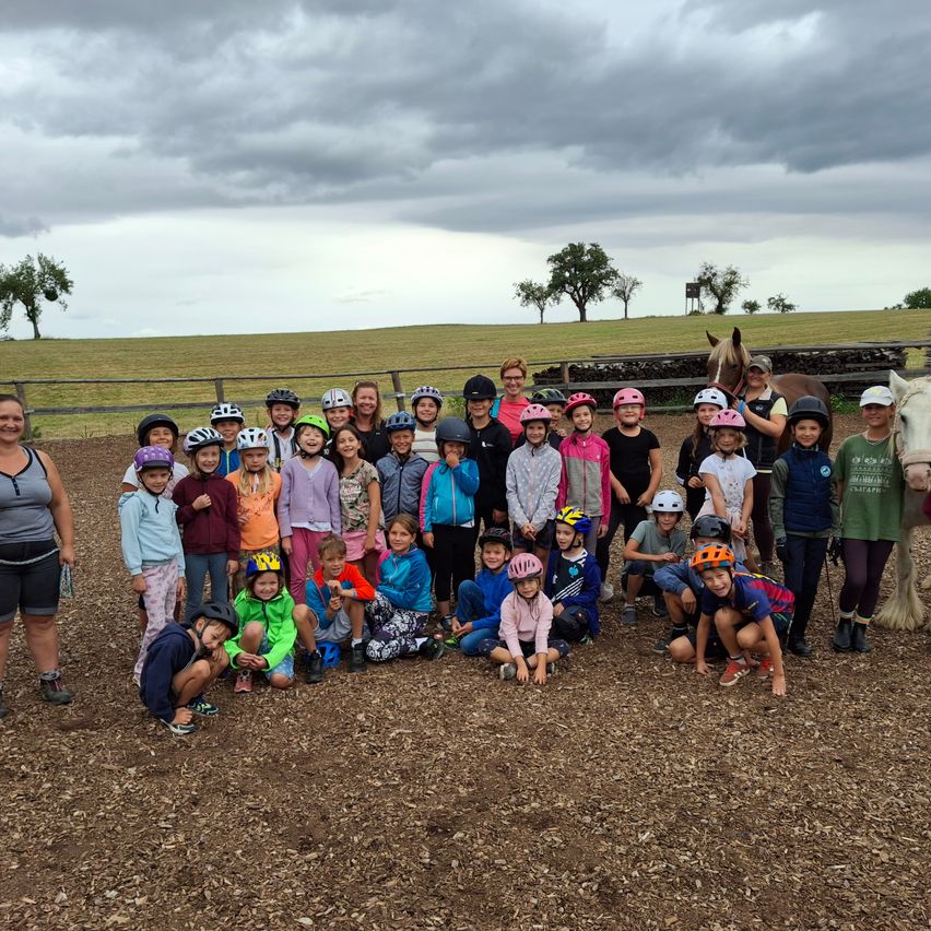 A group of children and adults wearing helmets stand in a dirt field with horses and a fence in the background.