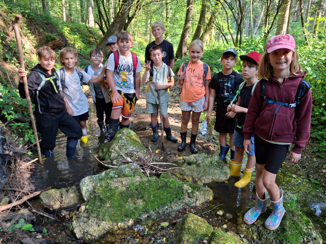 A group of children in a forest, some standing on rocks and others in water, all wearing boots and backpacks.