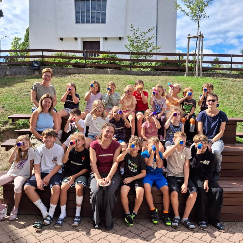 A group of children and adults are sitting on benches, holding up blue cups, possibly for a photo. They are in front of a white building with a fence.