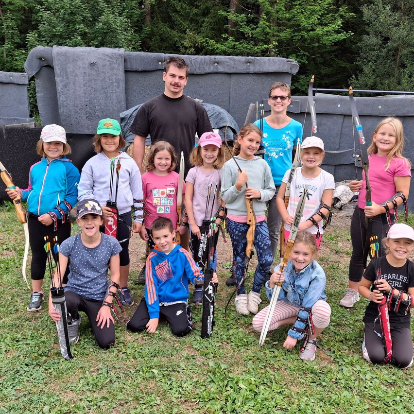 A group of children and adults are posing for a photo while holding bows and arrows. They are standing in a grassy area with a gray wall in the background.