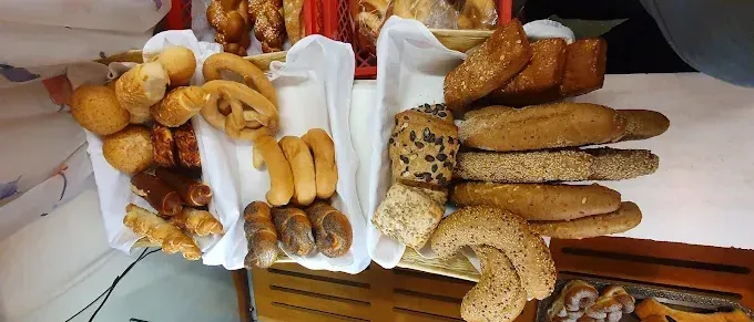 Eine Auswahl an Broten, darunter Croissants, Brezeln und Sesambrot, wird auf einer weißen Oberfläche präsentiert. Einige der Brote befinden sich in roten Körben, während andere auf einem Holztablett liegen.