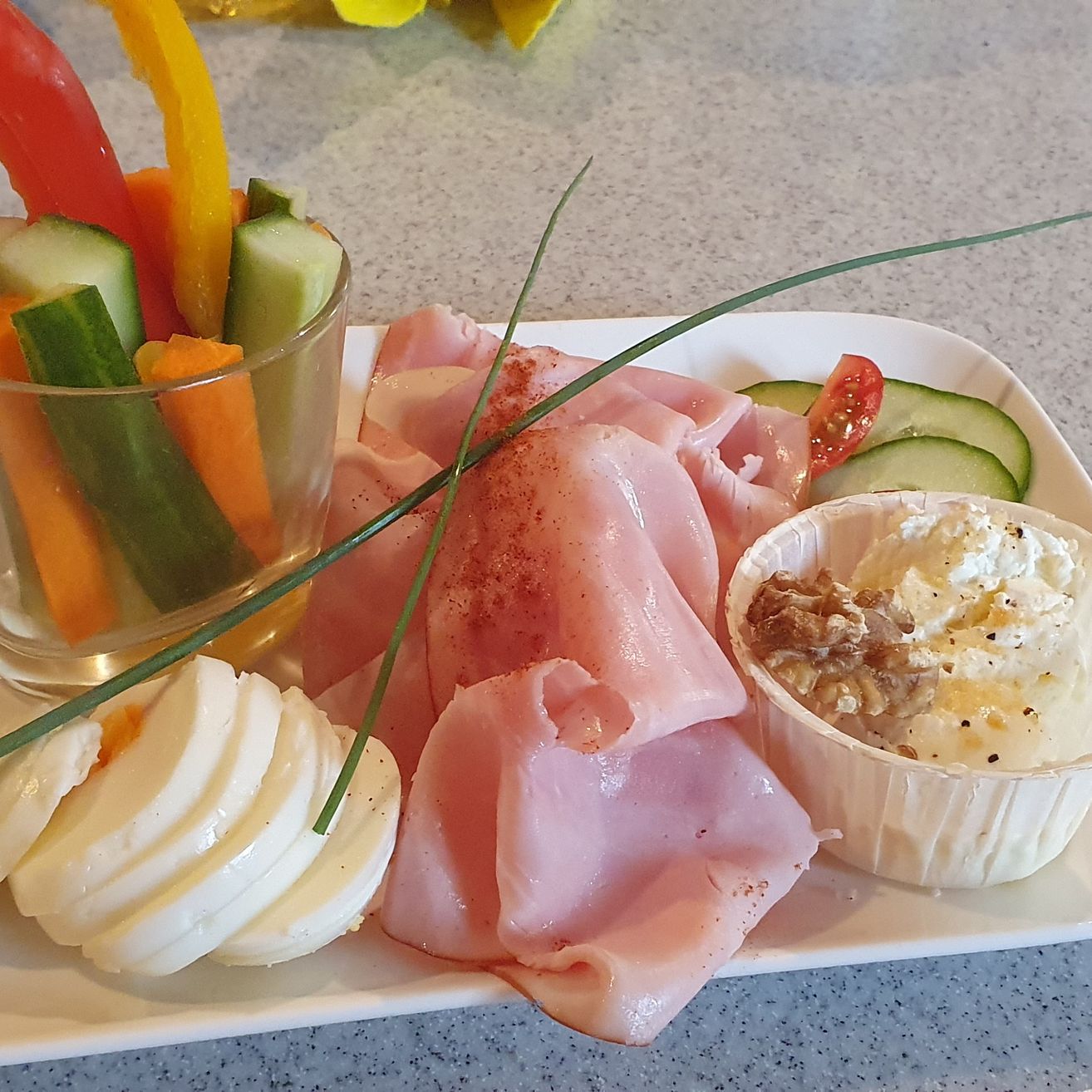 Ein weißer Teller hat geschnittenen Schinken, Gurken, Karotten und Paprika-Streifen und eine Schüssel mit Käse und Nüssen.