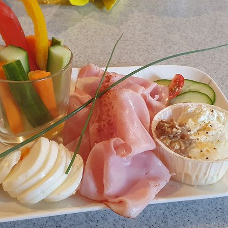 Ein weißer Teller hat geschnittenen Schinken, Gurken, Karotten und Paprika-Streifen und eine Schüssel mit Käse und Nüssen.