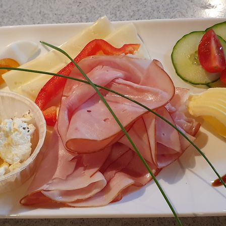 Ein weißer Teller enthält verschiedene Lebensmittel wie Schinkenscheiben, Käse, Gurken, Tomaten und rote Paprika, zusammen mit einer kleinen Schüssel Sahnekäse.