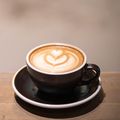 Eine Tasse Kaffee mit Latte-Kunst steht auf einem Untersetzer auf einem Holztisch. Der Hintergrund ist unscharf.