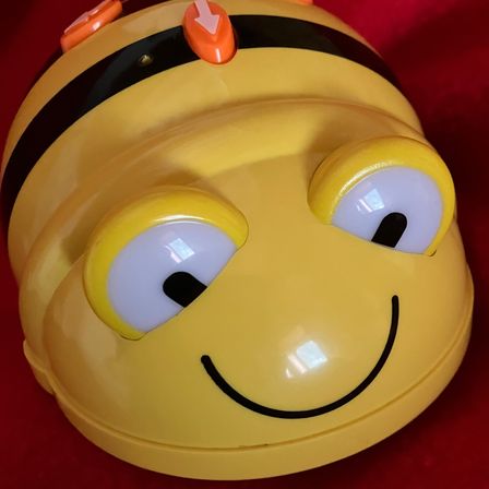 Ein gelbes Spielzeug, das eine Biene ähnelt, mit einem lächelnden Gesicht, schwarzen Antennen und orangefarbenen Knöpfen auf dem Kopf. Es hat weiße Augen mit schwarzen Pupillen.