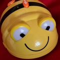 Ein gelbes Spielzeug, das eine Biene ähnelt, mit einem lächelnden Gesicht, schwarzen Antennen und orangefarbenen Knöpfen auf dem Kopf. Es hat weiße Augen mit schwarzen Pupillen.