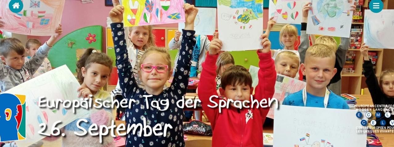 Kinder im Klassenzimmer halten Zeichnungen hoch mit den Texten 'September' und 'Tag der Sprachen'. Die Kinder lächeln und scheinen an einer Veranstaltung zum Tag der Sprachen teilzunehmen.