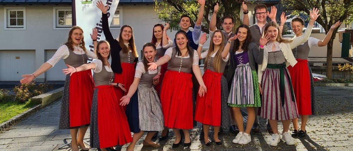 Eine Gruppe von Menschen in traditioneller Kleidung steht lächelnd vor einem Gebäude und einem Baum.