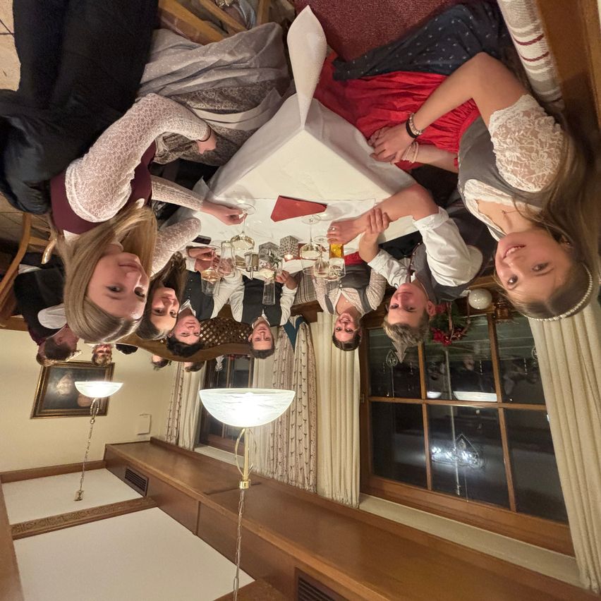 Eine Gruppe von Menschen versammelt sich um einen Tisch und lächelt für ein Foto. Der Tisch ist mit einer weißen Tischdecke bedeckt und hat Gläser und eine Kerze. Der Raum hat Holzbänke, Lampen und ein Fenster mit Vorhängen.