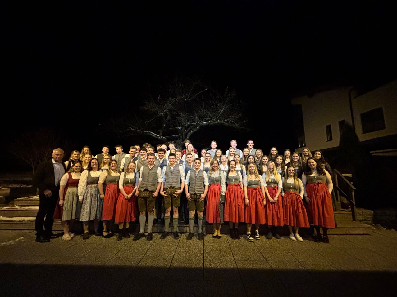 Eine Gruppe von Menschen in traditioneller Kleidung, möglicherweise für eine Aufführung, posiert für ein Foto bei Nacht vor einem Gebäude und einer Treppe.