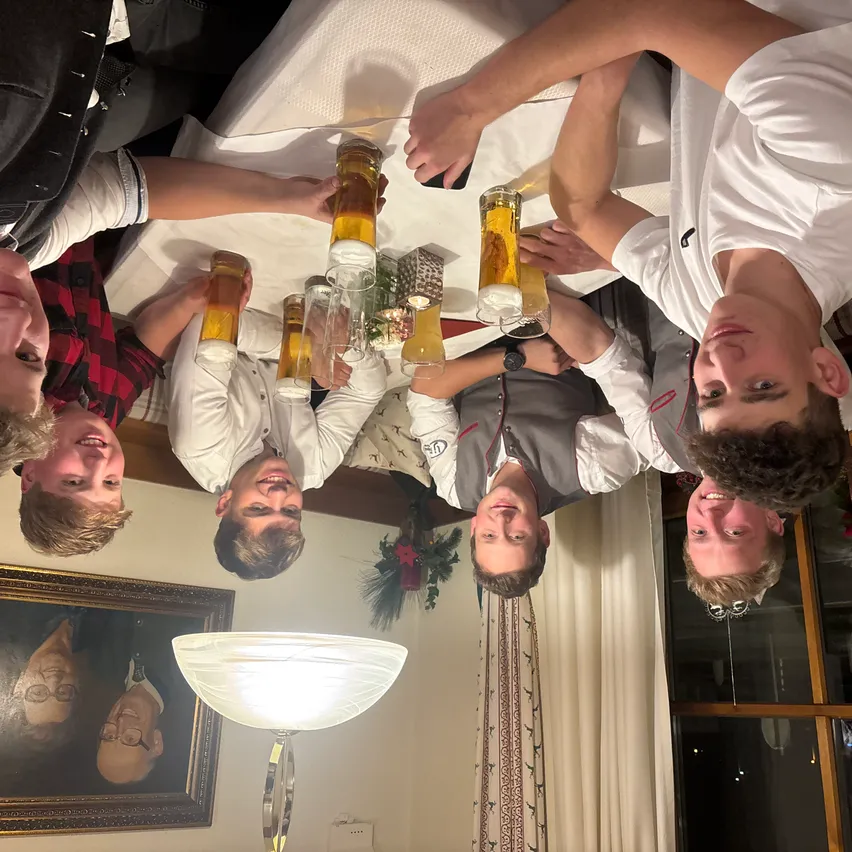 Eine Gruppe junger Männer lächelt und hält Gläser mit Bier. Sie sind kopfüber auf einem Tisch. Eine Lampe steht neben einem Bilderrahmen auf dem Boden.