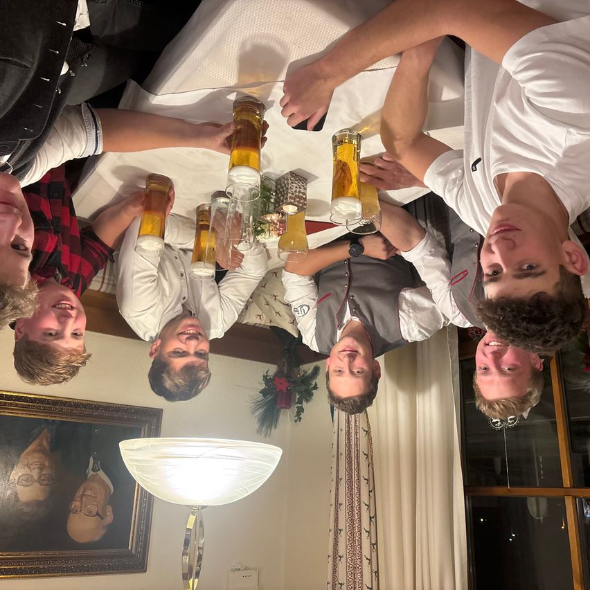 Eine Gruppe junger Männer lächelt und hält Gläser mit Bier. Sie sind kopfüber auf einem Tisch. Eine Lampe steht neben einem Bilderrahmen auf dem Boden.