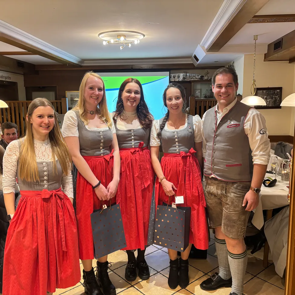 Fünf Menschen stehen eng zusammen für ein Foto. Sie tragen traditionelle Kleidung. Vier Frauen in roten Kleidern und ein Mann in traditioneller deutscher Kleidung. Sie halten schwarze Taschen mit roten Punkten. Dahinter befindet sich eine Projektionsleinwand.