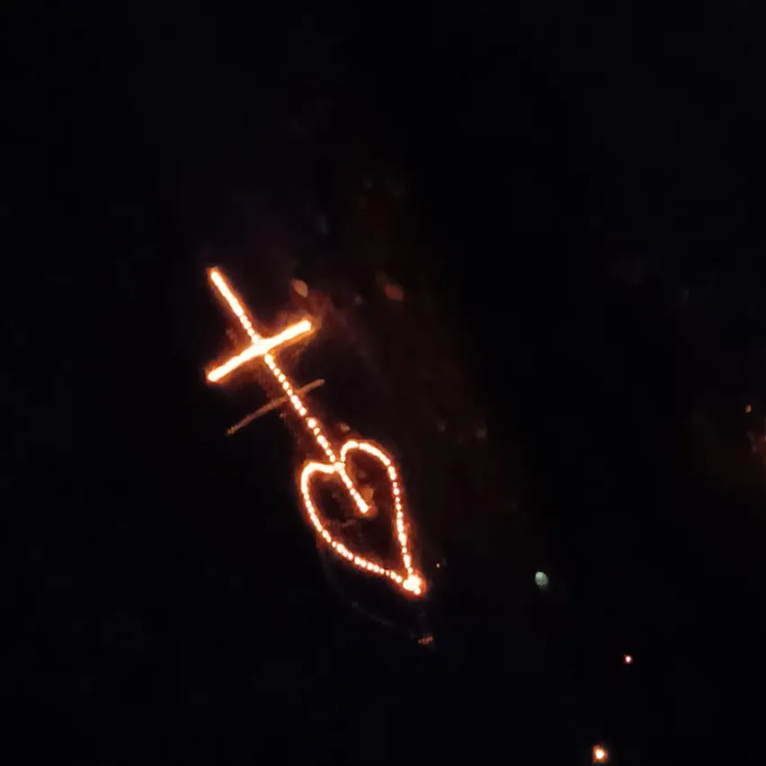 Ein Herzform mit einem Kreuz darüber, von einem warmen Licht beleuchtet, vor einem dunklen Hintergrund.