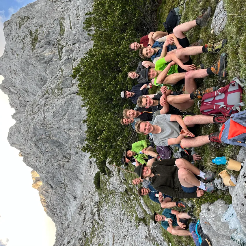 Eine Gruppe von Wanderern sitzt in einem Halbkreis am Berghang, einige mit Mützen und andere mit Stiefeln. Sie lächeln und posieren für ein Foto.