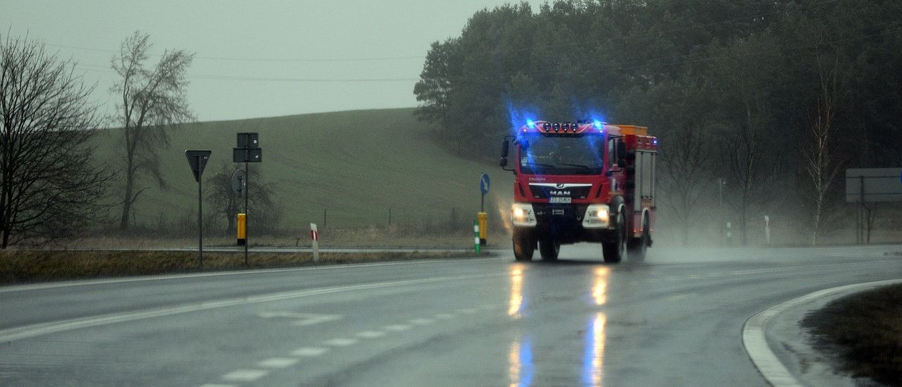 Ein roter Feuerwehrwagen fährt auf einer nassen Straße mit blinkenden blauen Lichtern, unter einem bewölkten Himmel mit Hintergrund von Bäumen.
