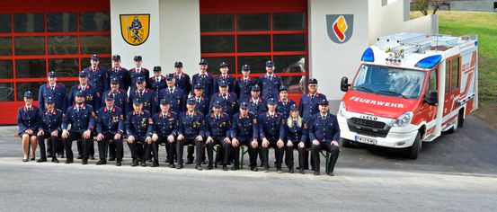 Eine Gruppe uniformierter Personen, möglicherweise Feuerwehrleute, posiert für ein Foto vor einer Feuerwache mit roten Türen und einem Logo. Ein Krankenwagen ist in der Nähe geparkt.