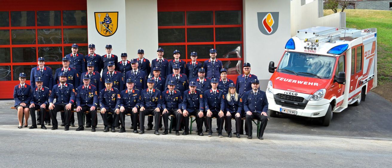 Eine Gruppe uniformierter Personen, möglicherweise Feuerwehrleute, posiert für ein Foto vor einer Feuerwache mit roten Türen und einem Logo. Ein Krankenwagen ist in der Nähe geparkt.