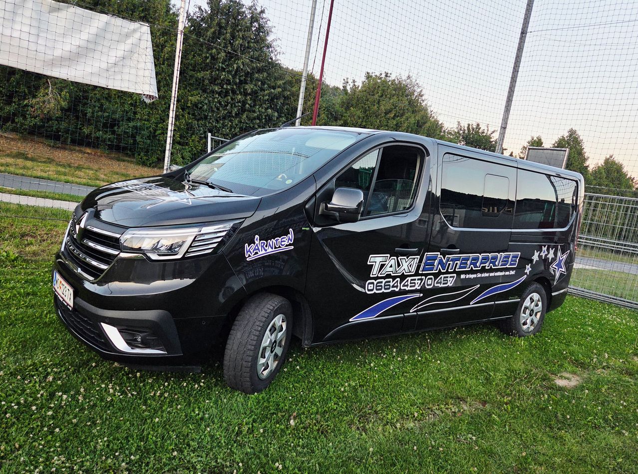 Ein schwarzer Minivan mit 'Kärnten' und 'TAXI ENTERPRISE' Aufklebern, geparkt auf einem Rasenfeld mit einem Zaun im Hintergrund.