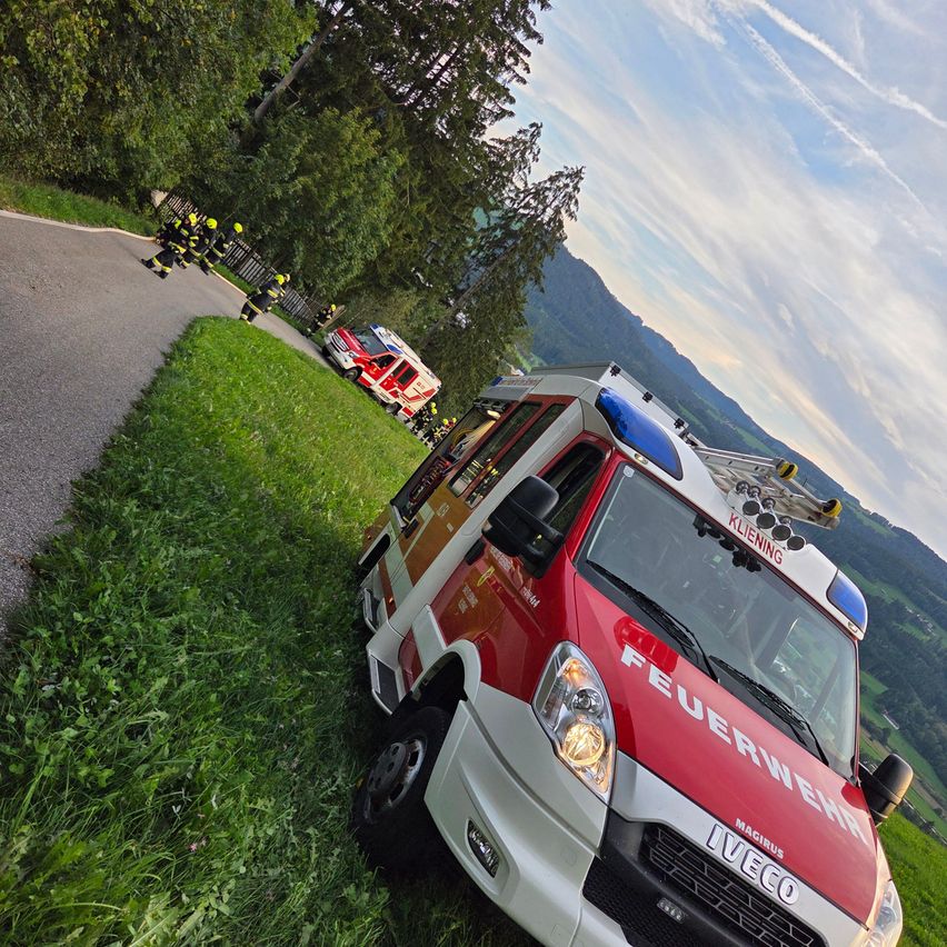 Ein rot-weißer Feuerwehrwagen steht am Straßenrand, Feuerwehrleute in gelben Helmen sind in der Nähe.