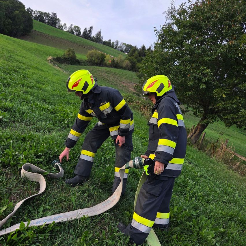 Zwei Feuerwehrleute in gelben Helmen und Uniformen arbeiten auf einem Grasfeld, einer hält eine Feuerwehrschläuche.