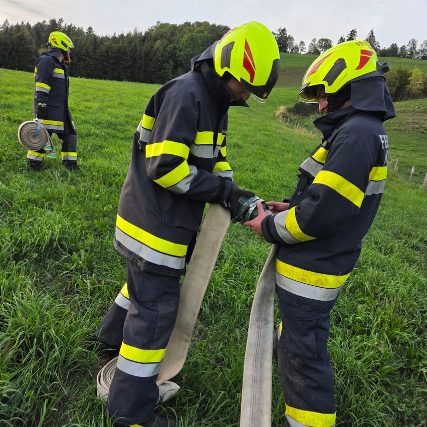 Drei Feuerwehrleute in voller Ausrüstung stehen auf einem Grasfeld, einer hält eine Feuerwehrschlauch und der andere eine Schlauchtrommel.