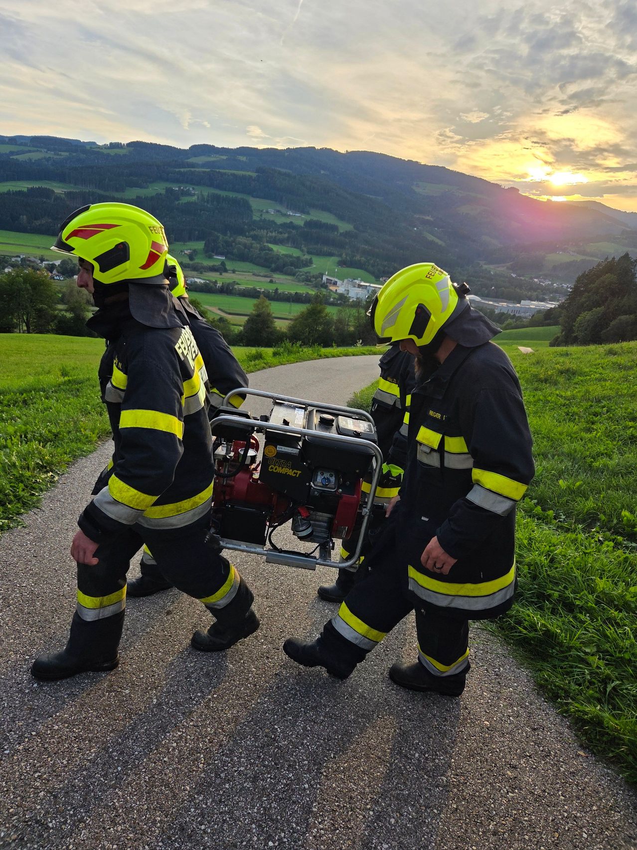 Drei Feuerwehrleute in gelben Helmen und schwarzen Uniformen tragen einen roten Generator auf einem Pfad durch ein Grasfeld mit Bergen im Hintergrund.
