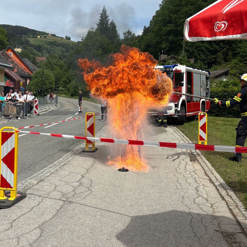Ein Feuerwehrmann löscht ein großes Feuer mit einem Schlauch auf einem offenen Gelände, mit Zuschauern an der Seite.