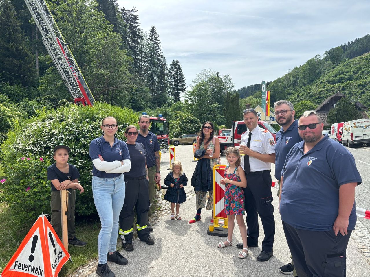 Eine Gruppe Feuerwehrleute und Zivilisten steht neben einem Feuerwehrwagen und einer Leiter, mit zwei Mädchen und einem Jungen davor und einem Mann, der ein Glas hält.