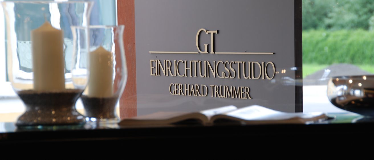 Ein Schild mit goldener Schrift, das 'GT Einrichtungsstudio Gerhard Trummer' zeigt, ist zu sehen. Eine Kerze und ein offenes Buch sind im Vordergrund.