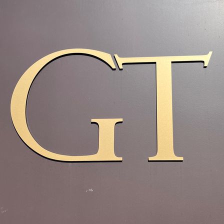 Ein braune Schild mit den goldenen Buchstaben GT ist auf einer rostigen Metalloberfläche montiert. Der Hintergrund ist ein weißes Gebäude mit einem Fenster.