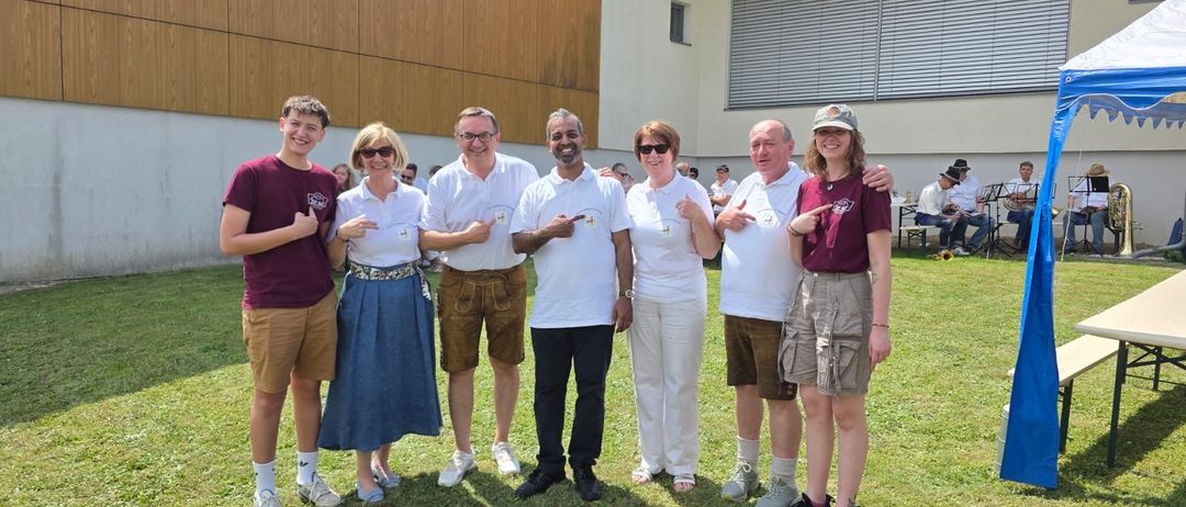 Eine Gruppe von Menschen in lässiger Kleidung steht vor einem Gebäude mit einem Grasfeld und sitzenden Musikern im Hintergrund.