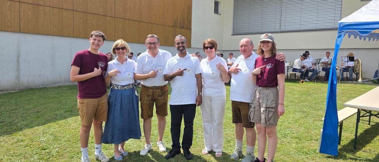 Eine Gruppe von Menschen in lässiger Kleidung steht vor einem Gebäude mit einem Grasfeld und sitzenden Musikern im Hintergrund.