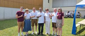 Eine Gruppe von Menschen in lässiger Kleidung steht vor einem Gebäude mit einem Grasfeld und sitzenden Musikern im Hintergrund.