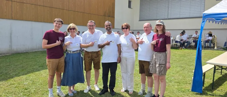 Eine Gruppe von Menschen in lässiger Kleidung steht vor einem Gebäude mit einem Grasfeld und sitzenden Musikern im Hintergrund.