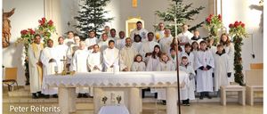 Eine Gruppe von Priestern und Kindern in weißen Gewändern posiert für ein Foto in einer Kirche, geschmückt mit Weihnachtsbäumen und Blumen.