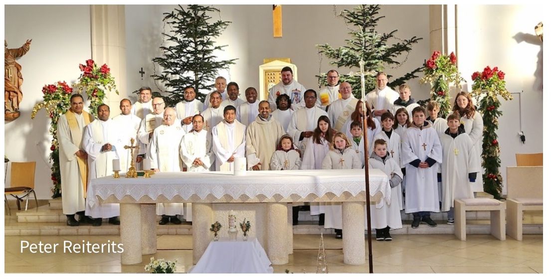 Eine Gruppe von Priestern und Kindern in weißen Gewändern posiert für ein Foto in einer Kirche, geschmückt mit Weihnachtsbäumen und Blumen.